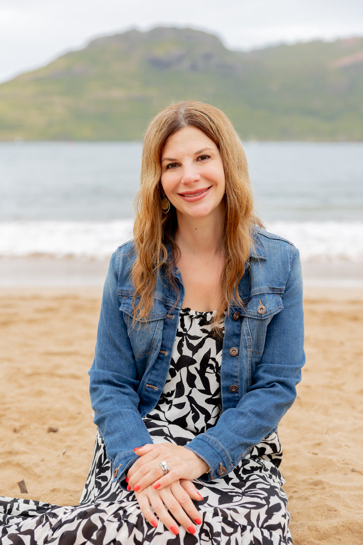 A woman with long light brown hair in a denim jacket and patterned dress smiles on a sandy beach with ocean views, enjoying seashell identification from *Seashells of Delmarva* by Schiffer Publishing, Ltd.