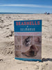 Seashells of Delmarva by Schiffer Publishing, Ltd. stands upright in the sand on a sunny beach, with the ocean in the background. The cover displays seashell photos, making it an ideal Delmarva seashell identification and beachcombing guide.
