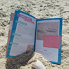 Seashells of Delmarva by Schiffer Publishing, Ltd. lies open on the sand beside a white shell, ocean, and blue sky—an ideal beachcombing guide for identifying and exploring Maryland and Virginia shells.