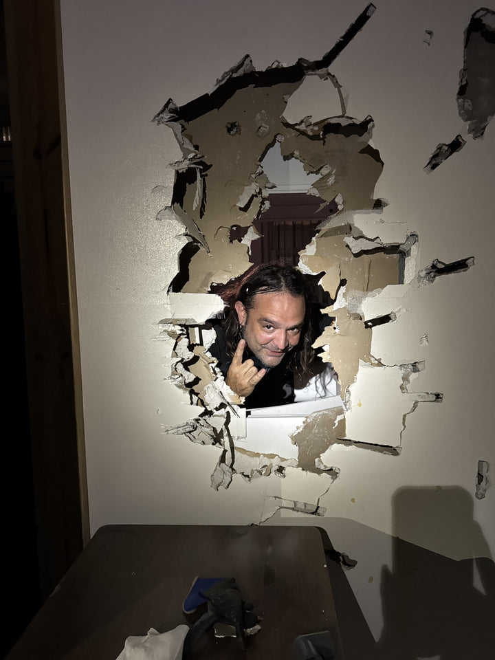 A man with long hair smiles and flashes a rock-and-roll hand sign through a hole in beige drywall, as if he’s just uncovered one of Savannah's mysteries from "Savannah's Afterlife III" by Schiffer Publishing, Ltd.