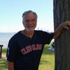 An older man in a navy blue Cleveland jersey smiles by a tree in a sunny lakeside park, with the city skyline and picnickers behind him—capturing an ideal moment for "Cleveland" by Schiffer Publishing, Ltd.