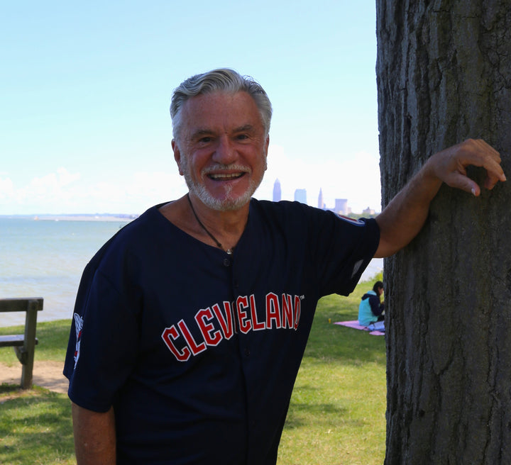 An older man in a navy blue Cleveland jersey smiles by a tree in a sunny lakeside park, with the city skyline and picnickers behind him—capturing an ideal moment for "Cleveland" by Schiffer Publishing, Ltd.