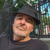 A man with a gray goatee, black fedora, and black shirt sits outdoors by an ornate iron fence and green trees, reflecting the artistic flair of graphic design France—perfect for fans of "The Little Book of French Signs" from Schiffer Publishing, Ltd.