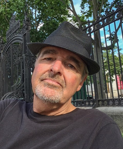 A man with a gray goatee, black fedora, and black shirt sits outdoors by an ornate iron fence and green trees, reflecting the artistic flair of graphic design France—perfect for fans of "The Little Book of French Signs" from Schiffer Publishing, Ltd.