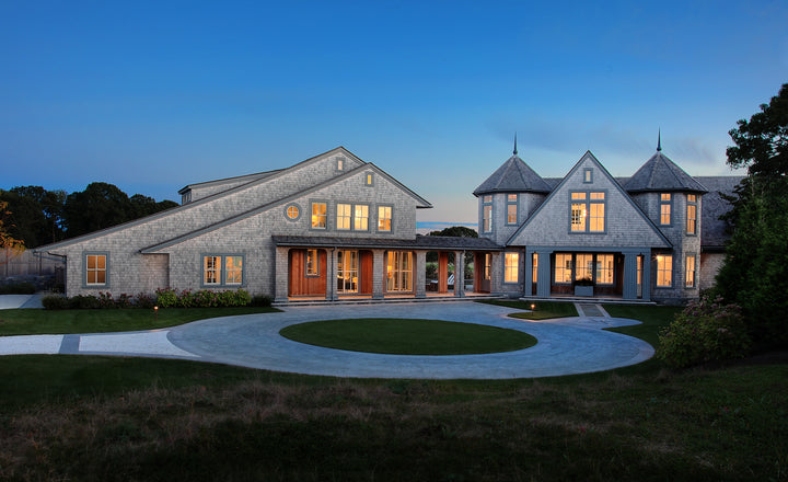 Shingle Style Houses by Schiffer Publishing, Ltd. features large modern gray homes with multiple peaked roofs, warmly lit windows, circular driveways, and manicured lawns highlighted at dusk.