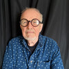 An older man with a gray beard and round glasses stands with arms crossed in a blue polka-dot shirt before a black curtain, embodying the spirit of "The Story of the Blues in 50 Songs" by Schiffer Publishing, Ltd.