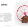 A woman in a white robe stands by a podium as art featured in "Keepers of a Movement" by Schiffer Publishing, Ltd.—like a vibrant pink bicycle wheel and house-shaped sculpture by Black artists—pops against the white background.