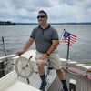 A man in sunglasses and casual attire steers a boat with an American flag on a cloudy day, capturing the spirit found in Schiffer Publishing, Ltd.'s "American Merchant Seaman's Manual, 8th Edition.