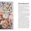 The book "Paper Flower Magic" by Schiffer Publishing, Ltd. rests beside pastel crepe-paper blooms in a glass vase, placed before an embroidered floral wall hanging and a book page titled Introduction about paper flowers.