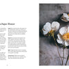 An open book, "Paper Flower Magic" by Schiffer Publishing, Ltd., displays instructions titled "Anatomy of a Paper Flower" on the left and a photo of three lifelike white and yellow crepe-paper flowers on a textured gray surface on the right.