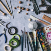 A flat lay of art and craft supplies for paper flower making, as featured in "Paper Flower Magic" by Schiffer Publishing, Ltd., including brushes, paints, scissors, pliers, tape, ruler, beads, clips, and skewers on a gray surface labeled "Tools.