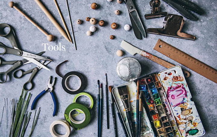 A flat lay of art and craft supplies for paper flower making, as featured in "Paper Flower Magic" by Schiffer Publishing, Ltd., including brushes, paints, scissors, pliers, tape, ruler, beads, clips, and skewers on a gray surface labeled "Tools.