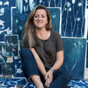 A woman with long brown hair sits barefoot in front of a wall displaying "Botanical Cyanotype" art prints from Schiffer Publishing, Ltd., smiling gently at the camera amidst the inspiring blue botanical artwork.