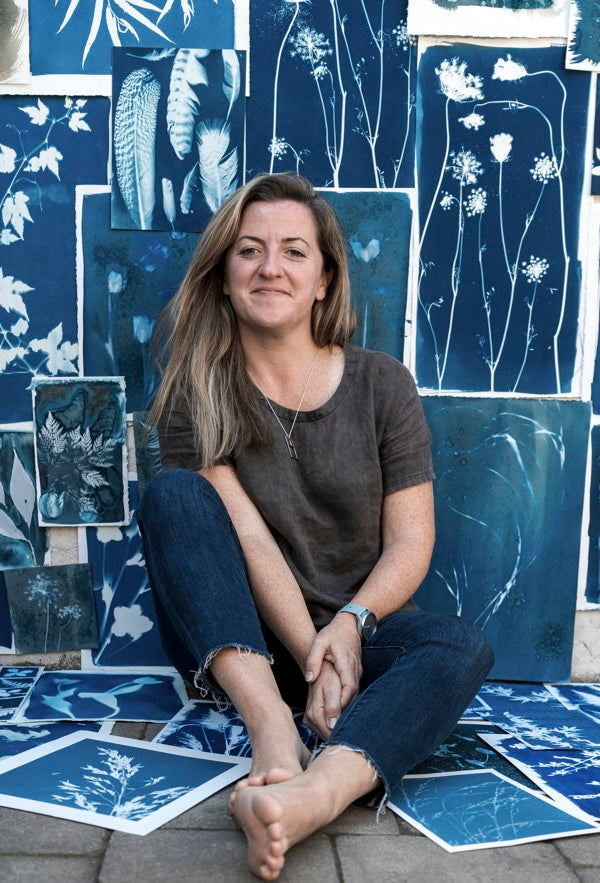 A woman with long brown hair sits barefoot in front of a wall displaying "Botanical Cyanotype" art prints from Schiffer Publishing, Ltd., smiling gently at the camera amidst the inspiring blue botanical artwork.