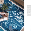 A person wraps a package on a table displaying the Botanical Cyanotype by Schiffer Publishing, Ltd., with white plant silhouettes on blue fabric. Ferns, twigs, a card, scissors, and twine spool are also arranged nearby.