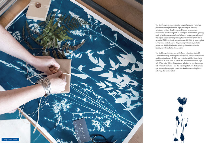 A person wraps a package on a table displaying the Botanical Cyanotype by Schiffer Publishing, Ltd., with white plant silhouettes on blue fabric. Ferns, twigs, a card, scissors, and twine spool are also arranged nearby.