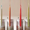 Four tall, slender candles—light beige, orange, red, and pale green—hang above metal molds with labels. Candle Whimsy by Schiffer Publishing, Ltd. is perfect for beginners or anyone exploring creative candle shapes.