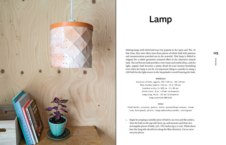 A wooden table with books and a small purple potted plant sits by a wood-paneled wall. Above, the Bark Craft lamp by Schiffer Publishing, Ltd. hangs from the ceiling, its birch bark weaving and leather trim adding charm to the cozy space.