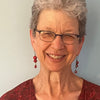 An older woman with short gray hair, glasses, and red earrings smiles at the camera in front of a light blue background. She wears a red patterned top reminiscent of "Tapestry Talking" by Schiffer Publishing, Ltd.