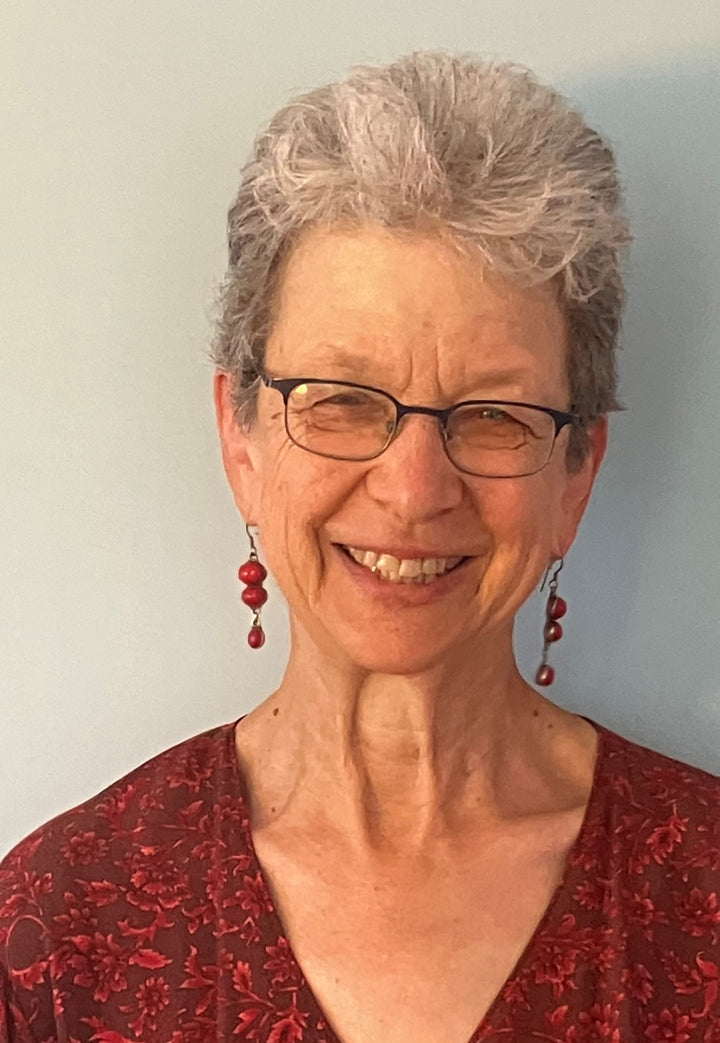 An older woman with short gray hair, glasses, and red earrings smiles at the camera in front of a light blue background. She wears a red patterned top reminiscent of "Tapestry Talking" by Schiffer Publishing, Ltd.