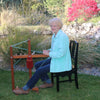An older woman with short blond hair and glasses sits outdoors by a pond, spinning yarn on a wheel for Nordic projects, as featured in The Bandweaver's Project Book from Schiffer Publishing, Ltd.