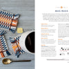 Two handwoven mug rugs in blue, orange, white, and beige stripes sit on a wooden surface near a cup of coffee. In the foreground is a third rug and metal spoon. "The Bandweaver's Project Book" by Schiffer Publishing, Ltd. appears at right.