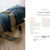 The Bandweaver's Project Book by Schiffer Publishing, Ltd. is pictured on a wooden surface next to a rolled blanket secured with a yellow and gray inkle-loom woven strap, complete with metal buckles, handles, and printed sling carrier instructions.