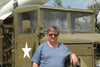 A man in sunglasses and a blue t-shirt stands by an olive green military truck with a missile, evoking the iconic Sd.Kfz. vehicles featured in Schiffer Publishing, Ltd.'s German Light Armored Cars of WWII.