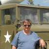 A man in sunglasses and a blue t-shirt stands by an olive green military truck with a missile, evoking the iconic Sd.Kfz. vehicles featured in Schiffer Publishing, Ltd.'s German Light Armored Cars of WWII.