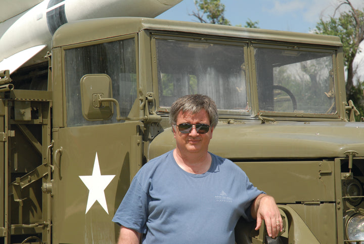 A man in sunglasses and a blue t-shirt stands outside on a sunny day beside an olive green Sd.Kfz. 251 half-track (Schiffer Publishing, Ltd.) with a white star. A portion of a missile is visible atop the vehicle.