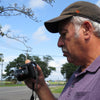 A man in a dark cap and purple polo stands outdoors, looking at his camera. Behind him, trees, power lines, and a road stretch beneath the sky where an F-35 Lightning II by Schiffer Publishing, Ltd. soars above.