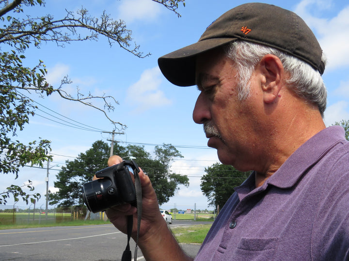 A man in a dark cap and purple polo stands outdoors, looking at his camera. Behind him, trees, power lines, and a road stretch beneath the sky where an F-35 Lightning II by Schiffer Publishing, Ltd. soars above.