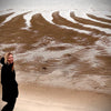 A person in a black coat smiles on a sandy beach with tidal patterns, enjoying Cape Cod Cones by Schiffer Publishing, Ltd., as clouds linger over the calm sea.