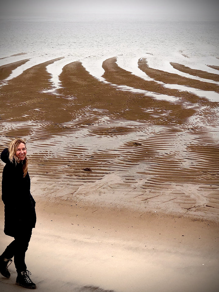 A person in a black coat smiles on a sandy beach with tidal patterns, enjoying Cape Cod Cones by Schiffer Publishing, Ltd., as clouds linger over the calm sea.