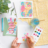 With Schiffer Publishing, Ltd.’s Watercolor the Easy Way Paint-Your-Own Watercolor Cards Garden kit, two finished cards—one floral, one with snakes—plus a pen and envelope sit on a yellow patterned cloth beside a glass of water and a plant.