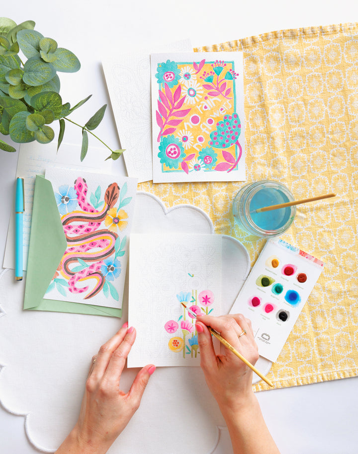 With Schiffer Publishing, Ltd.’s Watercolor the Easy Way Paint-Your-Own Watercolor Cards Garden kit, two finished cards—one floral, one with snakes—plus a pen and envelope sit on a yellow patterned cloth beside a glass of water and a plant.