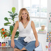 A smiling blonde woman in a white sleeveless top and blue jeans sits cross-legged on a wooden table, surrounded by plants and creating with Schiffer Publishing, Ltd.’s Watercolor the Easy Way Paint-Your-Own Watercolor Cards Friends.