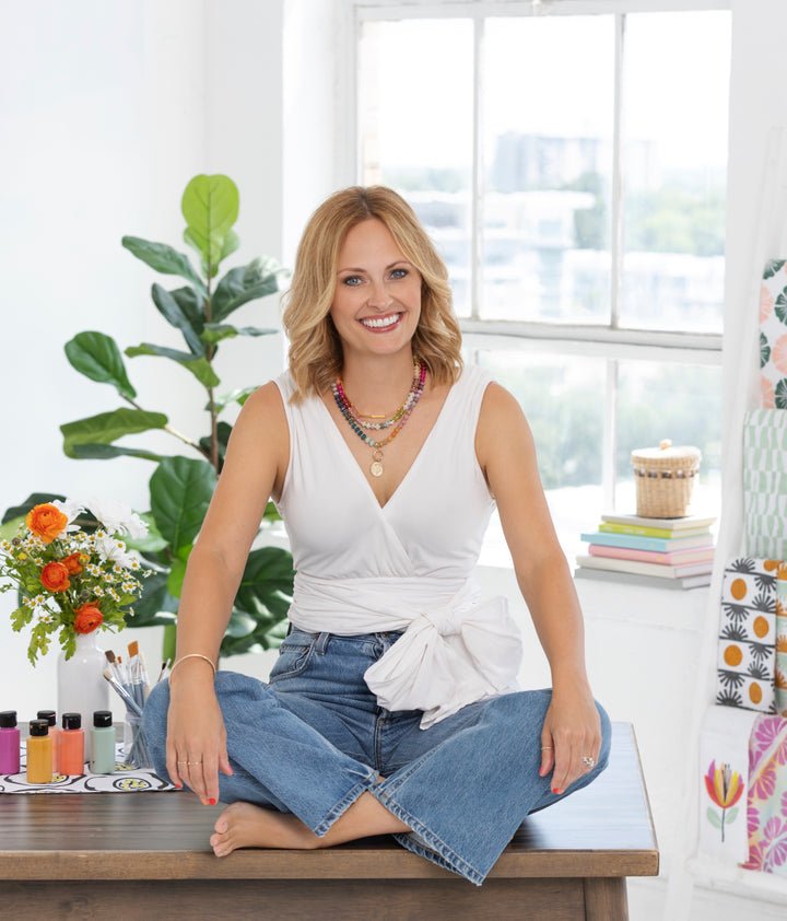 A smiling blonde woman in a white sleeveless top and blue jeans sits cross-legged on a wooden table, surrounded by plants and creating with Schiffer Publishing, Ltd.’s Watercolor the Easy Way Paint-Your-Own Watercolor Cards Friends.