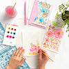 Using the Watercolor the Easy Way Paint-Your-Own Watercolor Cards Friends set by Schiffer Publishing, Ltd., a person paints floral cards at a table with paint, brushes, a pink drink, finished cards, a pink pen, an envelope, and a plant.