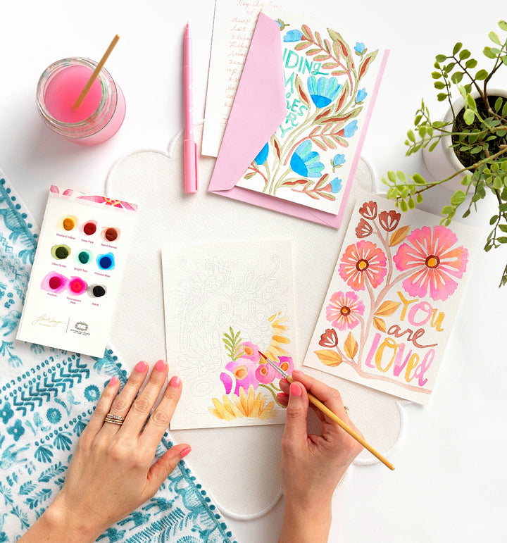 Using the Watercolor the Easy Way Paint-Your-Own Watercolor Cards Friends set by Schiffer Publishing, Ltd., a person paints floral cards at a table with paint, brushes, a pink drink, finished cards, a pink pen, an envelope, and a plant.