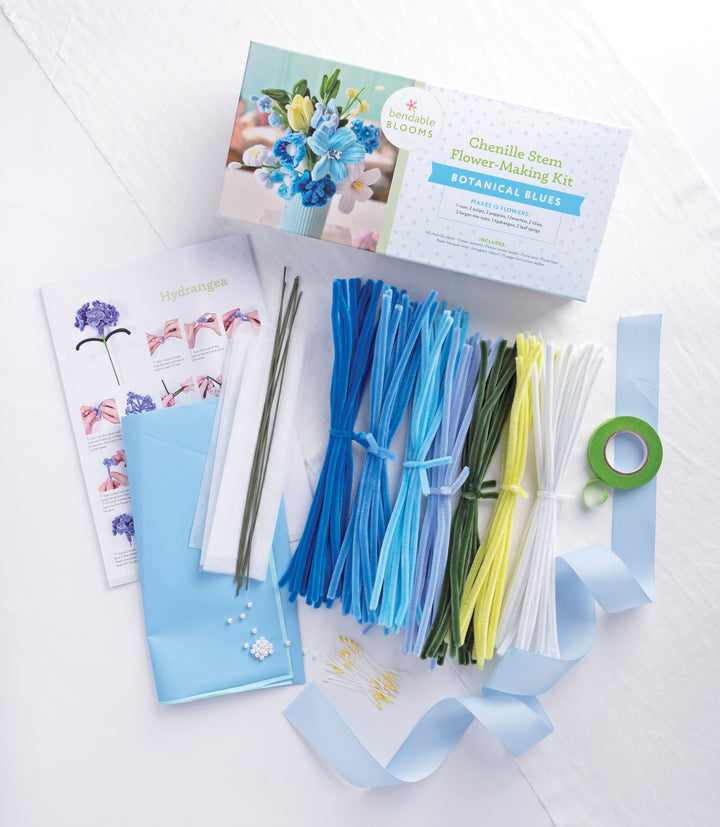 The "Bendable Blooms Chenille Stem Flower-Making Kit Botanical Blues" by Schiffer Publishing, Ltd. includes blue, white, and yellow pipe cleaners, floral tape, wire stems, beads, hydrangea instructions, and a blue envelope on a white backdrop.