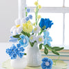 A white vase and small bowl display a DIY bouquet made with Schiffer Publishing, Ltd.'s Bendable Blooms Chenille Stem Flower-Making Kit Botanical Blues on a light green mat near books and a window.