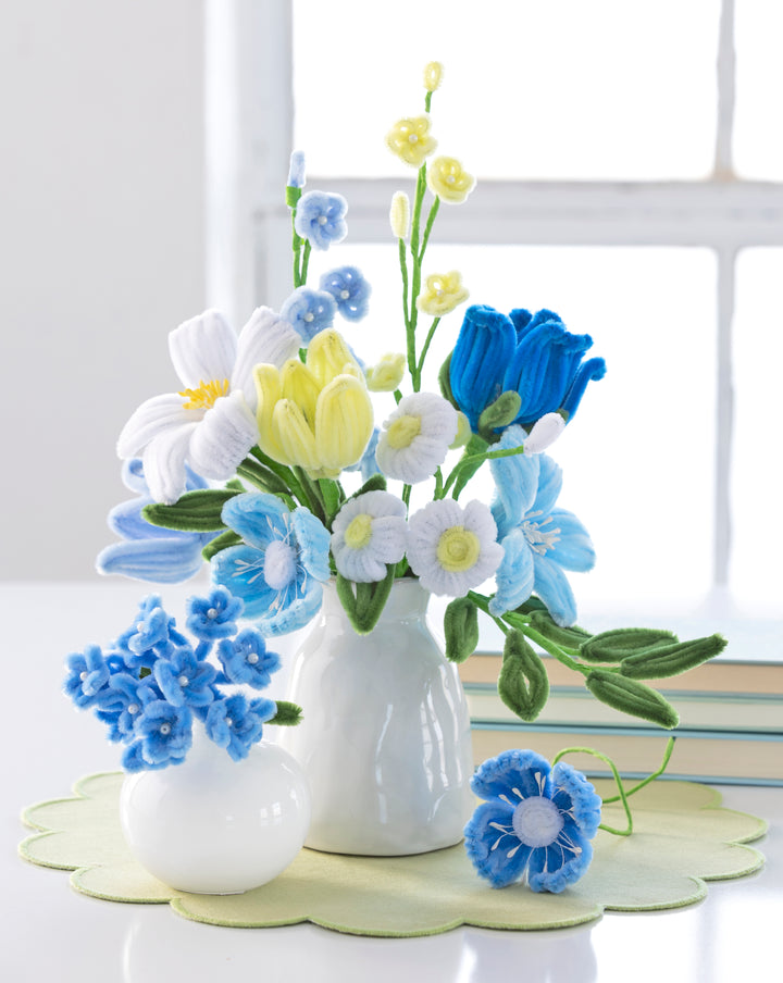 A white vase and small bowl display a DIY bouquet made with Schiffer Publishing, Ltd.'s Bendable Blooms Chenille Stem Flower-Making Kit Botanical Blues on a light green mat near books and a window.