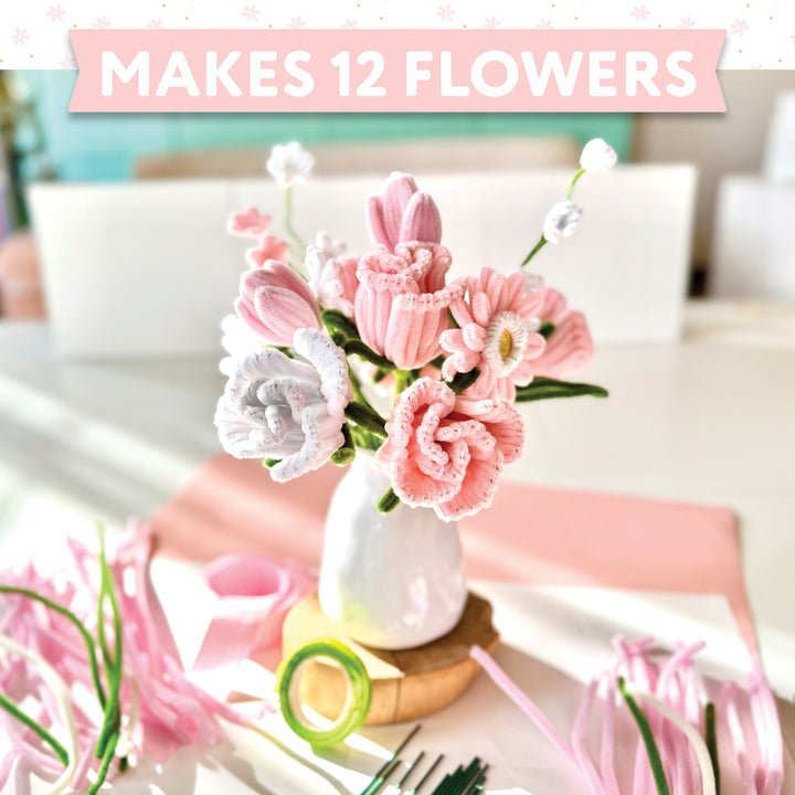 A vase with 12 handmade pink and white flowers sits on a table with craft supplies from the Schiffer Publishing, Ltd. Bendable Blooms Chenille Stem Flower-Making Kit Cottage Garden. A banner at the top reads MAKES 12 FLOWERS.