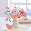 A white vase displays pink and white felt flowers made with the Bendable Blooms Chenille Stem Flower-Making Kit Cottage Garden by Schiffer Publishing, Ltd. Nearby is a pink bird ornament on a yellow placemat, with books and a dish by the window.