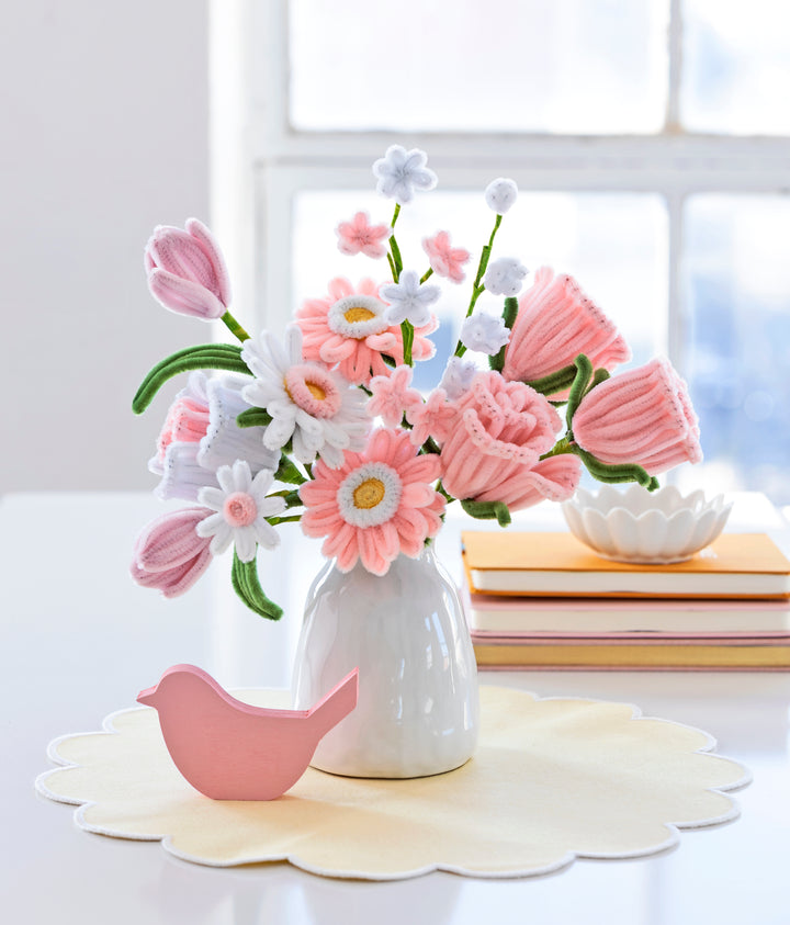 A white vase displays pink and white felt flowers made with the Bendable Blooms Chenille Stem Flower-Making Kit Cottage Garden by Schiffer Publishing, Ltd. Nearby is a pink bird ornament on a yellow placemat, with books and a dish by the window.