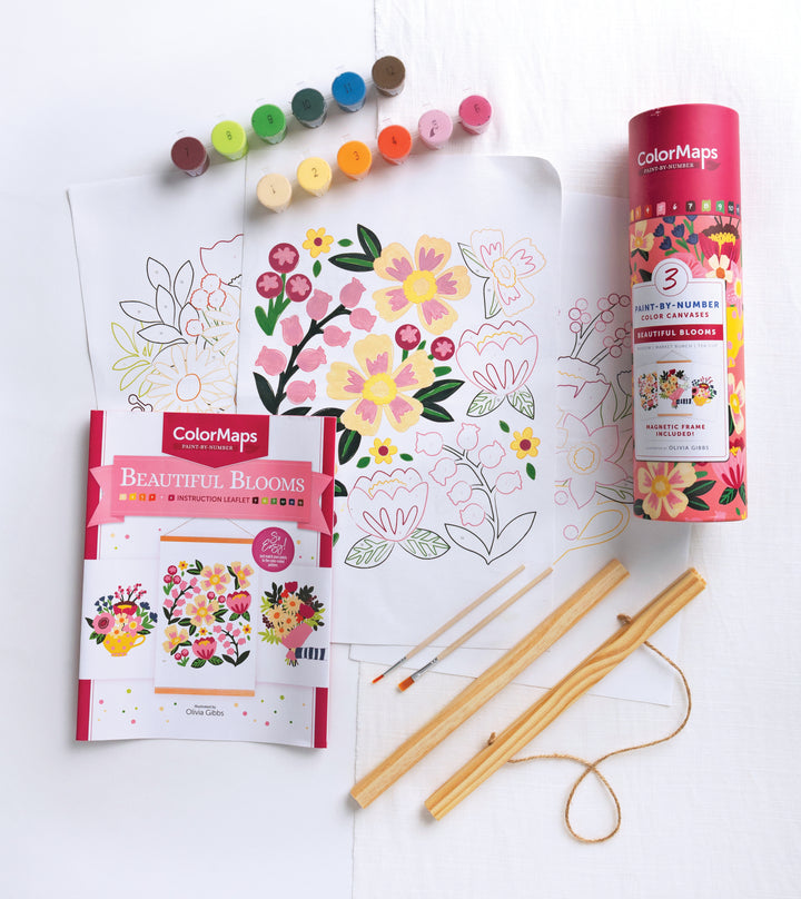 The ColorMaps Paint-by-Number Beautiful Blooms kit by Schiffer Publishing, Ltd. sits on a table with paint pots, two brushes, a vibrant floral outline sheet, folded instructions, and a pink cylindrical ColorMaps container.