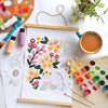 The ColorMaps Paint-by-Number Beautiful Blooms by Schiffer Publishing, Ltd. is in progress on white paper, surrounded by paintbrushes, watercolor paints, bottles, a palette, coffee cup, plant, and another floral sketch.