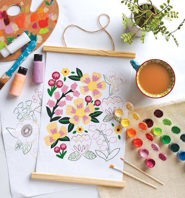 The ColorMaps Paint-by-Number Beautiful Blooms by Schiffer Publishing, Ltd. is in progress on white paper, surrounded by paintbrushes, watercolor paints, bottles, a palette, coffee cup, plant, and another floral sketch.
