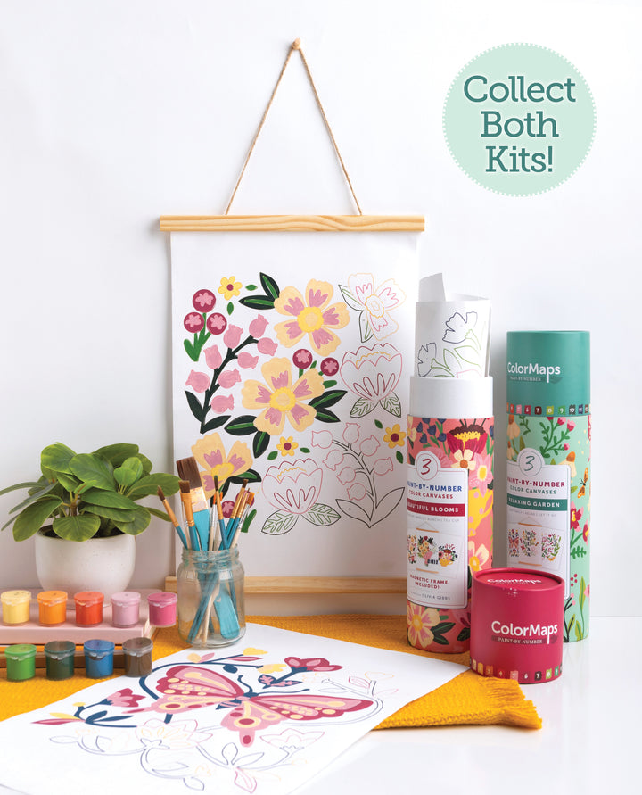 The ColorMaps Paint-by-Number Beautiful Blooms kit by Schiffer Publishing, Ltd. includes paint pots, brushes in a jar, a small plant, and rolled kit tubes. A completed artwork hangs above with a “Collect Both Kits!” circle—great for beginner painters.